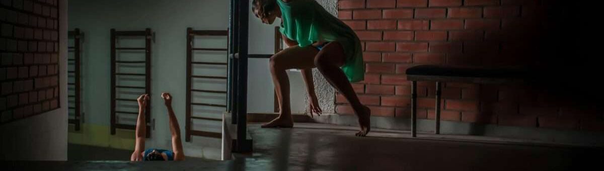 A woman dances expressively indoors with dramatic lighting against a brick wall in Santa Cruz, Bolivia.